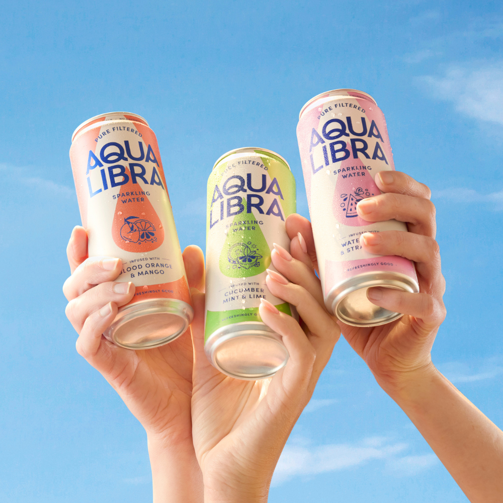Three cans of Aqua Libra sparkling water held by hands against a blue sky.